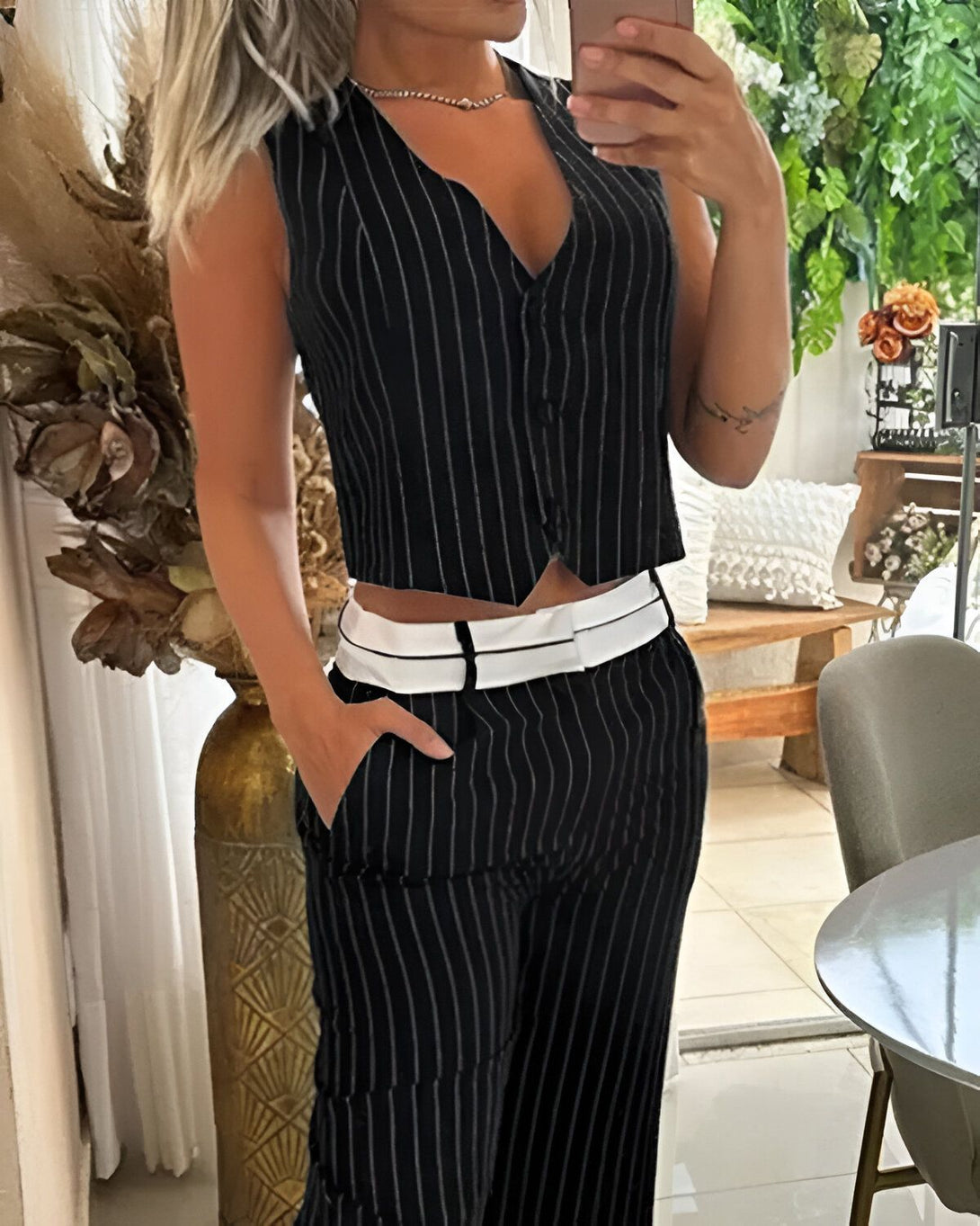 Une femme porte un gilet noir et blanc à rayures fines et un pantalon noir et blanc à rayures fines. Elle a une main dans la poche du pantalon et l'autre tient un téléphone portable.