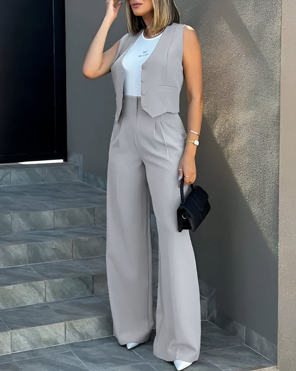 Une femme porte un tailleur pantalon gris avec un gilet et un pantalon large. Elle porte également un sac à main noir et des escarpins blancs.