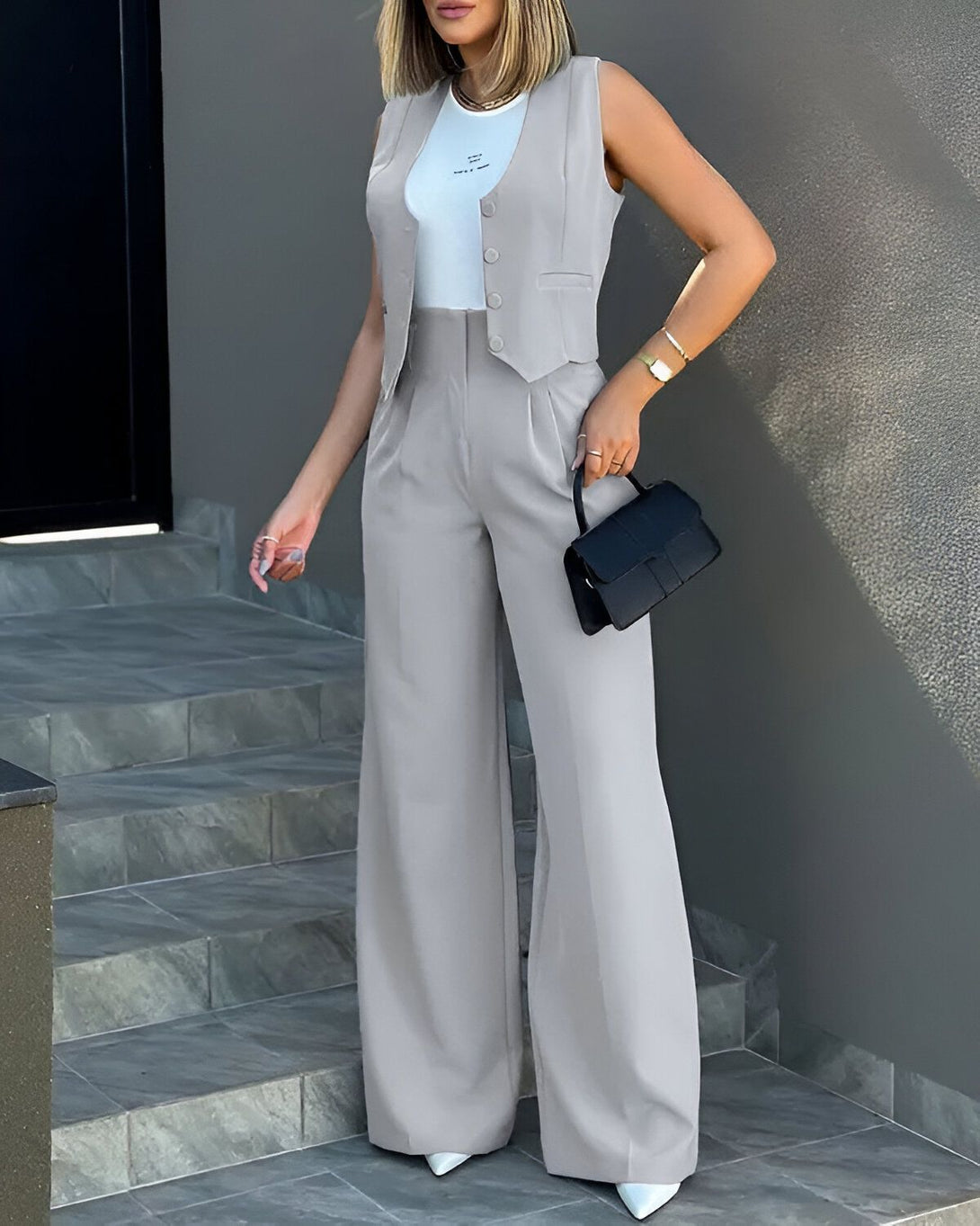 Une femme porte un gilet gris et un pantalon gris. Elle porte également un sac à main noir et des talons blancs.