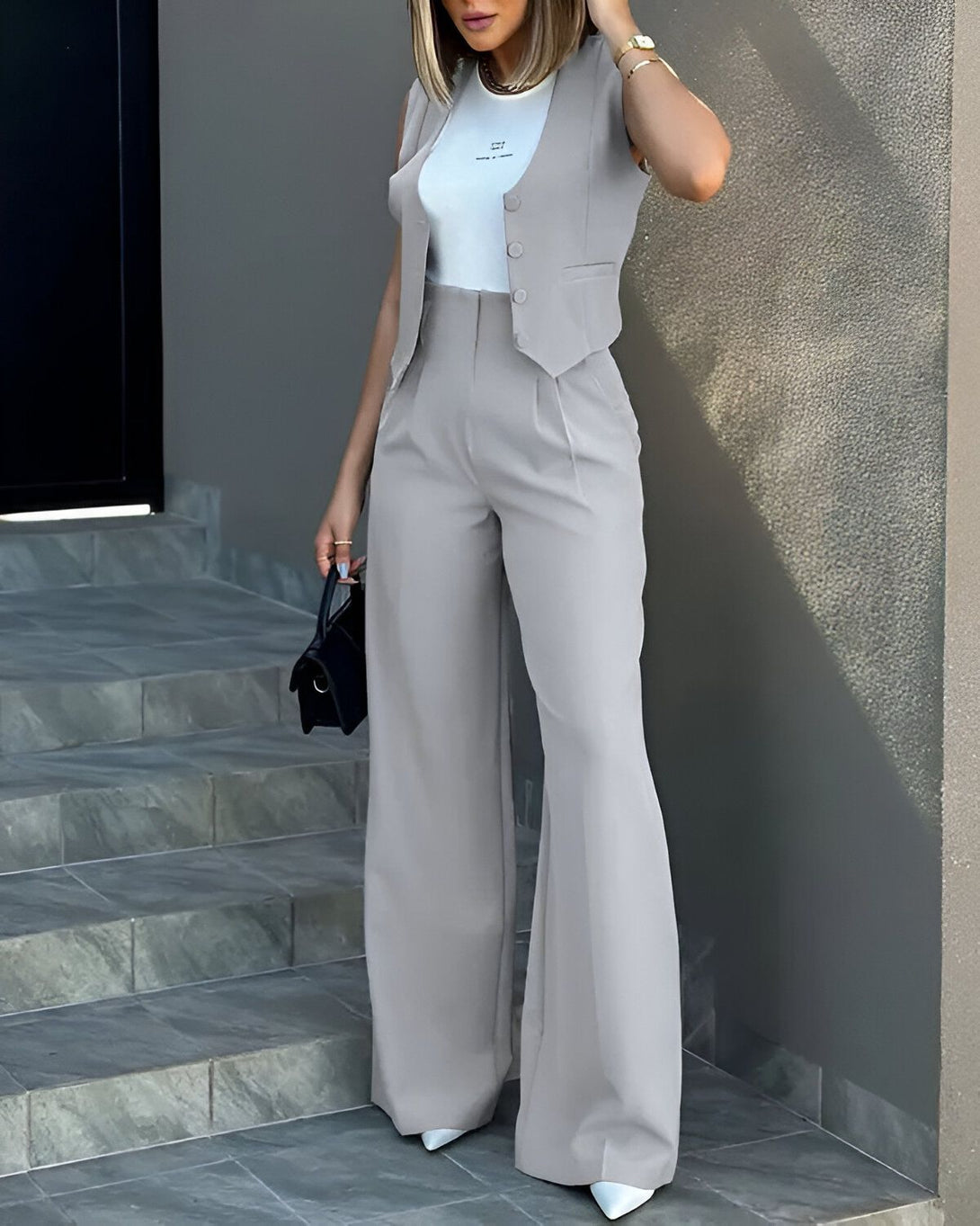 Une femme porte un tailleur pantalon gris avec un gilet et un pantalon large. Elle porte également un sac à main noir et des escarpins blancs.