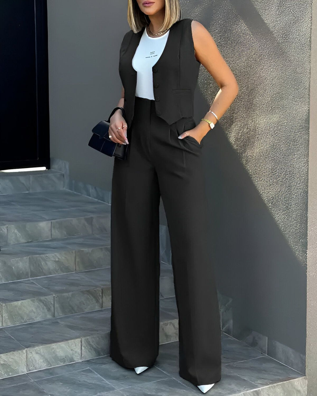 Une femme porte un gilet noir et un pantalon large noir. Elle porte également un sac à main noir et des talons blancs.
