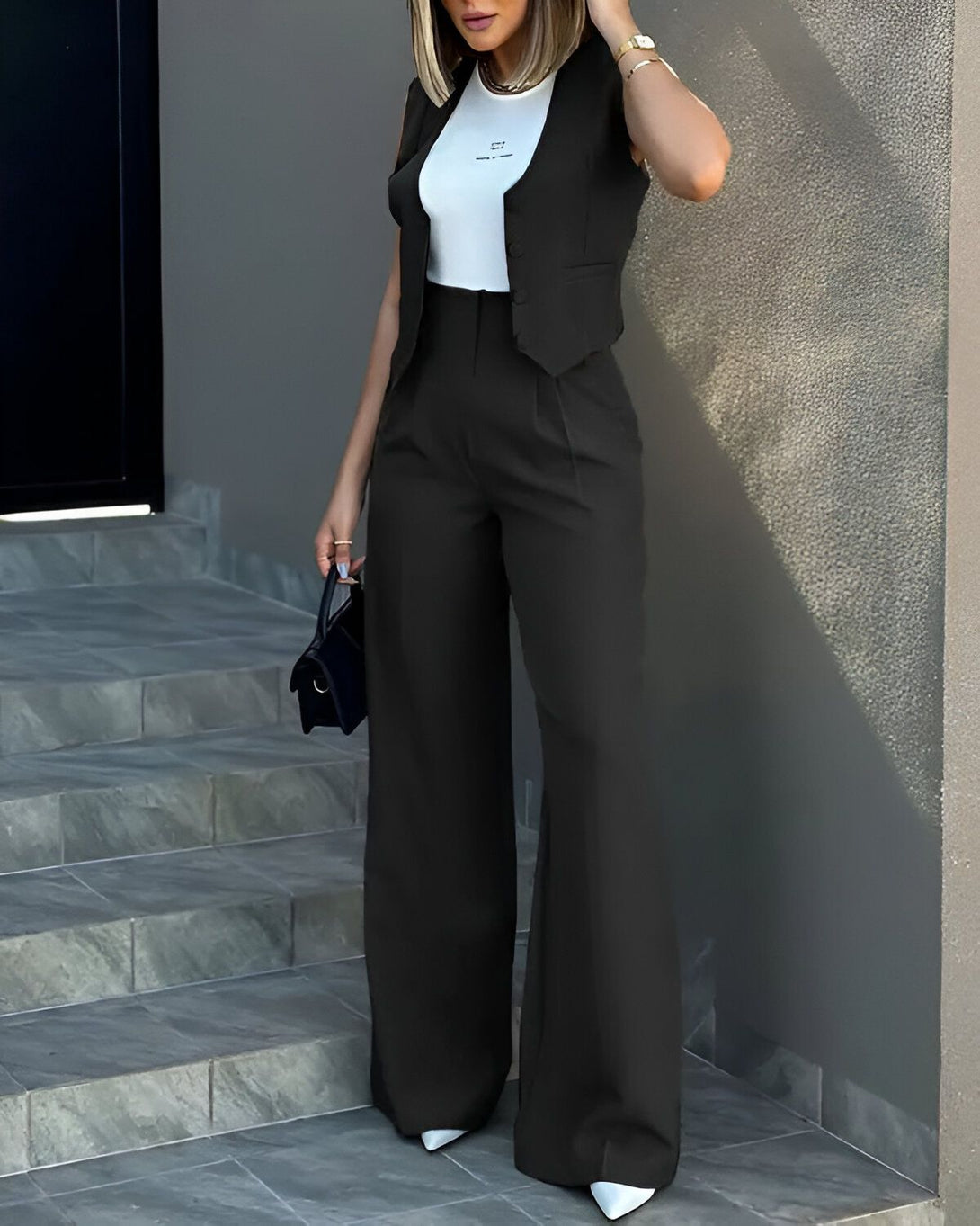 Une femme porte un tailleur pantalon noir et un gilet noir. Elle porte également un sac à main noir et des escarpins blancs.