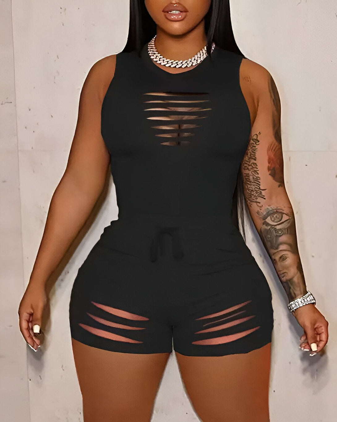 Une femme porte un ensemble deux pièces noir. Le haut est un débardeur court avec des déchirures au niveau de la poitrine. Le short est taille haute et possède également des déchirures.