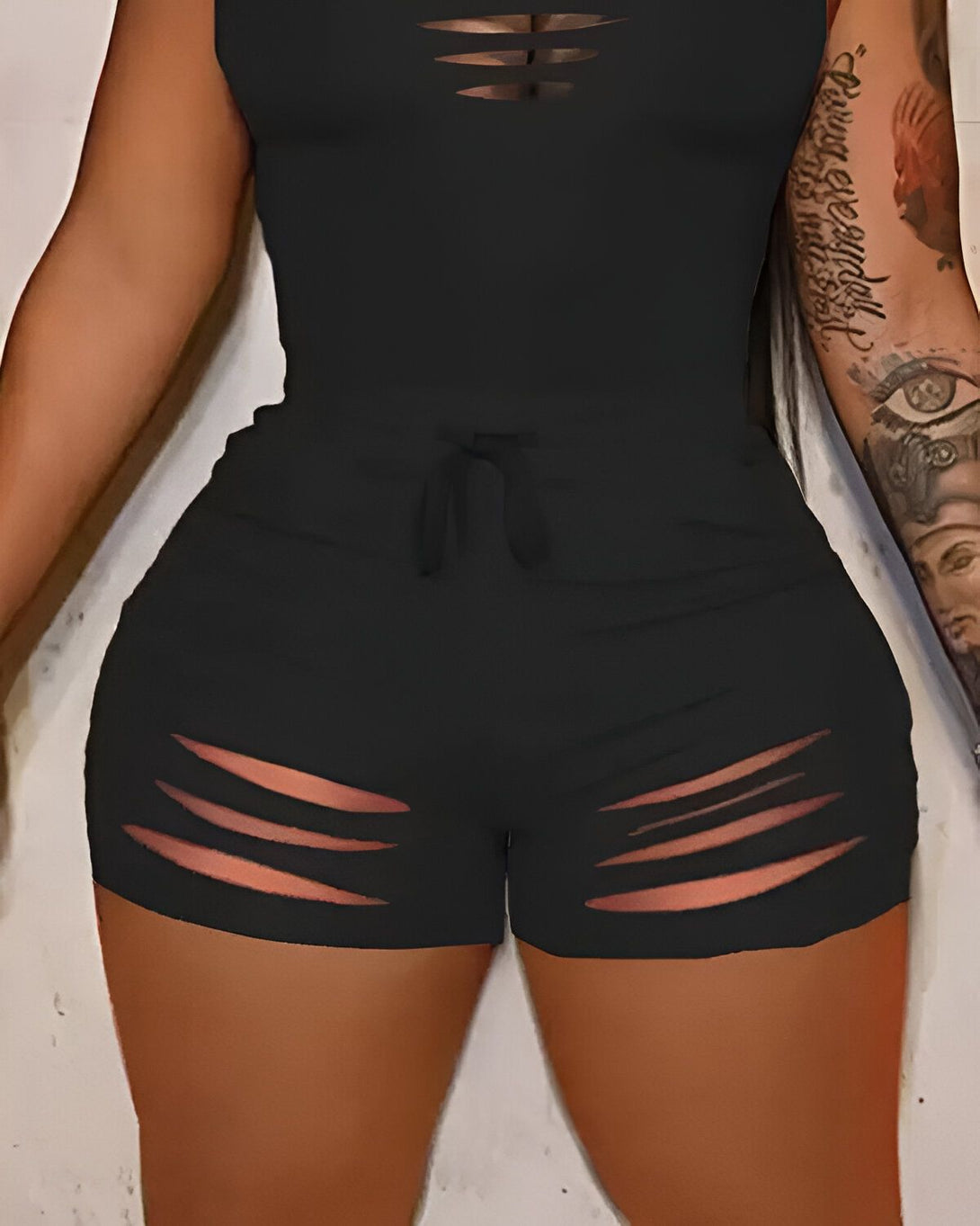 Une femme porte un ensemble deux pièces noir. Le haut est un débardeur court avec des déchirures au niveau de la poitrine. Le short est taille haute et possède également des déchirures au niveau des cuisses.