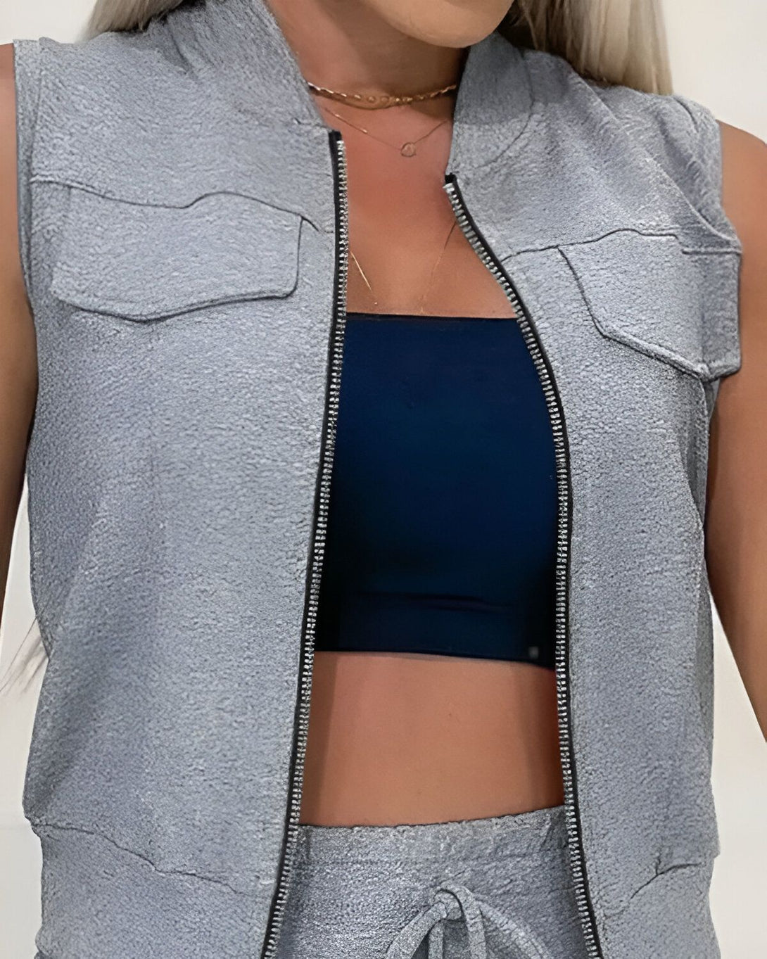 Une femme porte un gilet gris sans manches et un pantalon de jogging gris assorti. Elle porte également un soutien-gorge bleu marine.