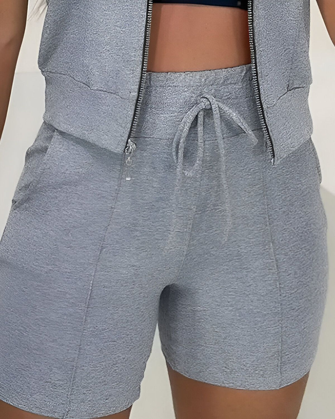 Une femme porte un short gris taille haute avec une veste zippée assortie. Le short est doté d'une coulisse à la taille et de poches latérales.
