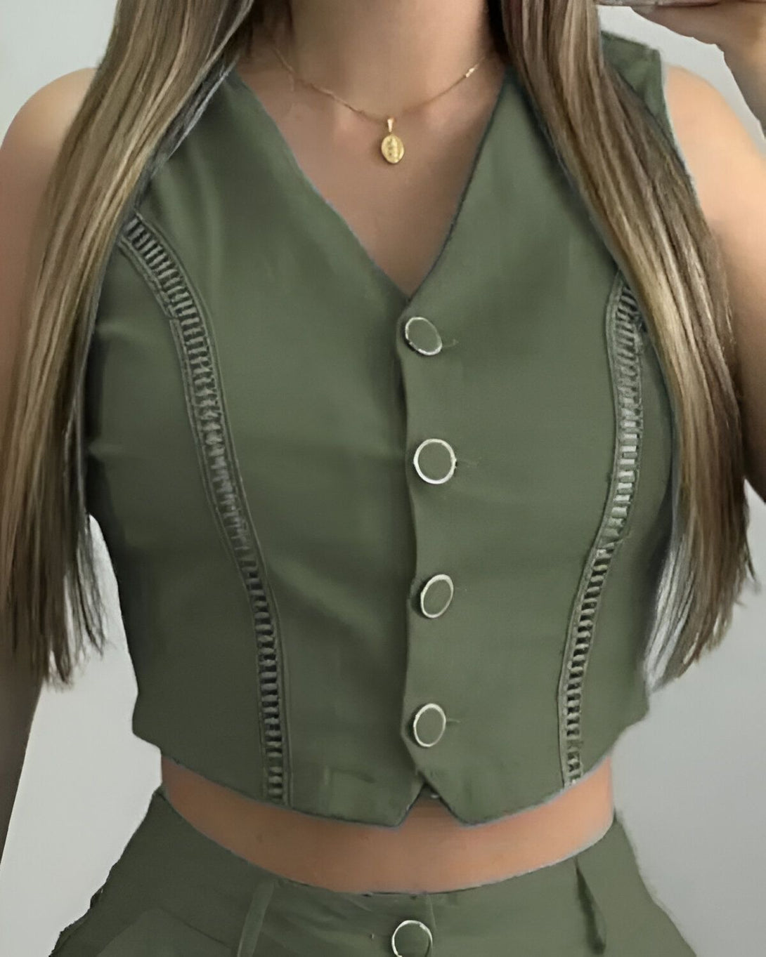 Une jeune femme porte un gilet vert olive et un pantalon assorti. Le gilet est court et cintré, avec des boutons sur le devant. Le pantalon est taille haute et évasé. La femme a les cheveux longs et blonds, et elle porte un collier avec un pendentif rond.