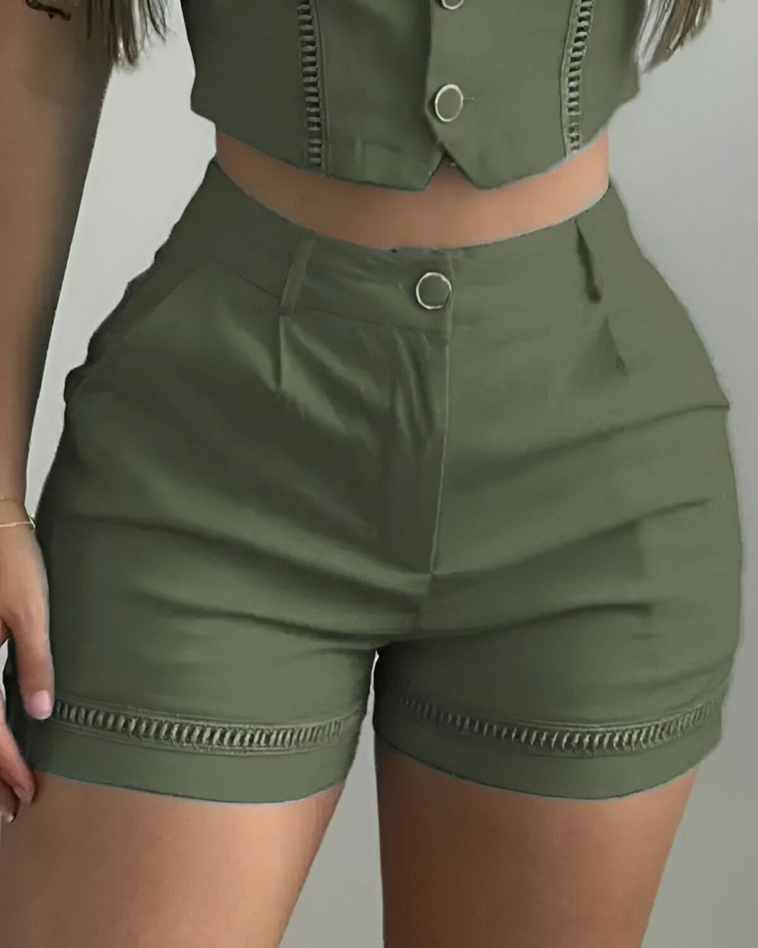 Une femme porte un short taille haute vert olive avec des boutons dorés. Le short est cintré et possède des poches sur les côtés. Il est porté avec un haut court blanc.