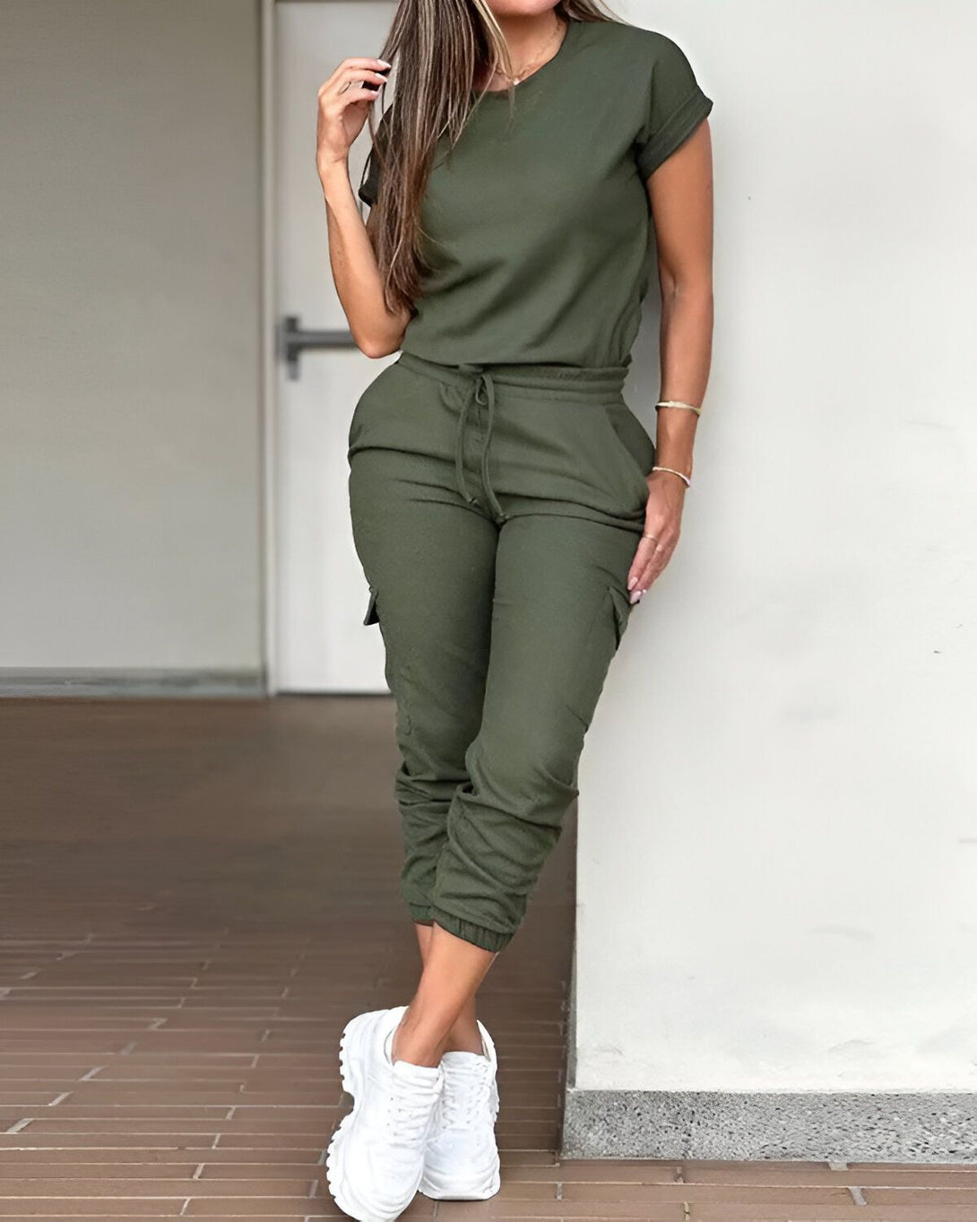 Une femme porte un ensemble vert kaki composé d'un t-shirt à manches courtes et d'un pantalon cargo. Elle est debout, adossée à un mur, et porte des baskets blanches.