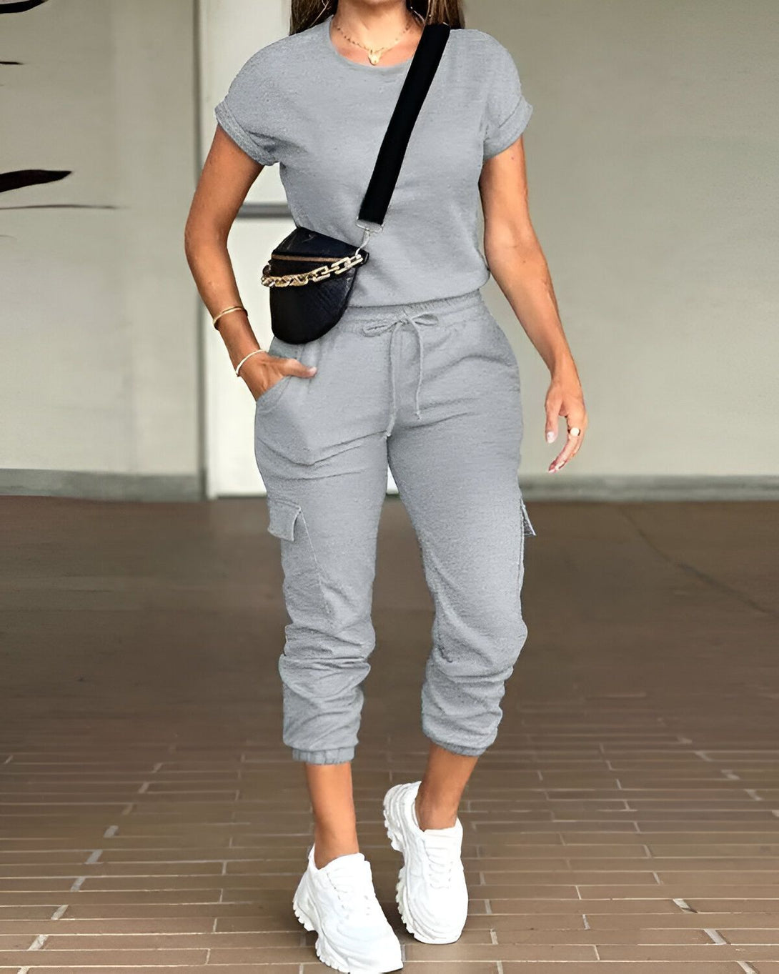 Une femme porte un ensemble jogging gris composé d'un t-shirt à manches courtes et d'un pantalon cargo. Elle porte également des baskets blanches et un sac banane noir.