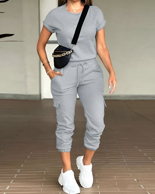 Une femme porte un ensemble jogging gris composé d'un t-shirt à manches courtes et d'un pantalon cargo. Elle porte également des baskets blanches et un sac banane noir.