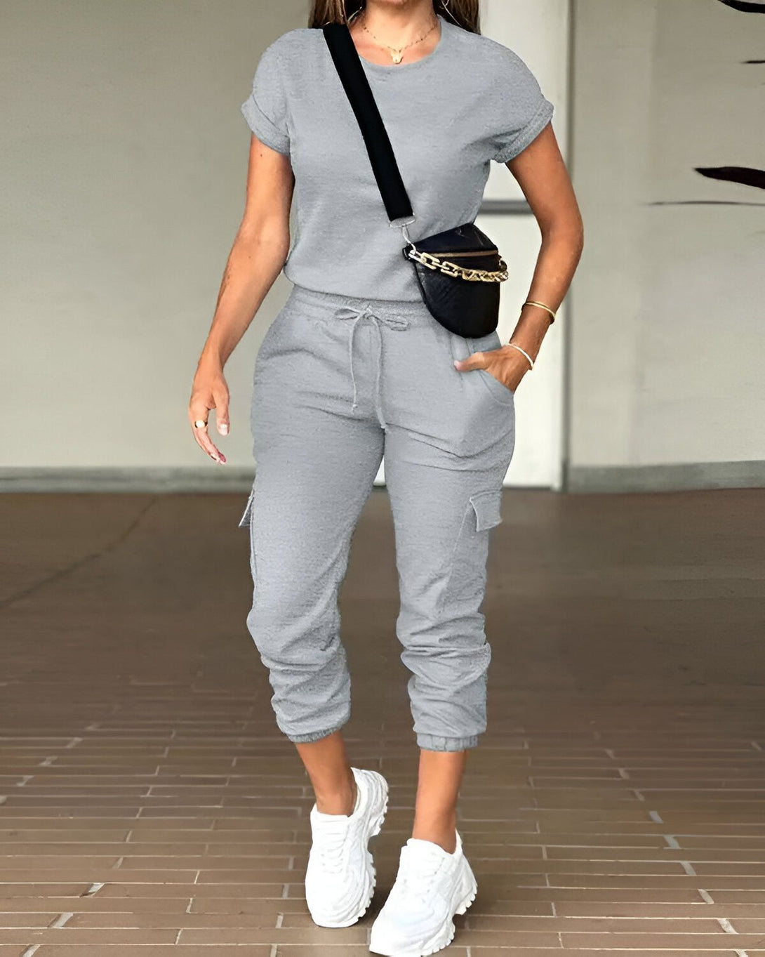 Une femme porte un ensemble jogging gris composé d'un t-shirt à manches courtes et d'un pantalon cargo. Elle porte également des baskets blanches et un sac banane noir.