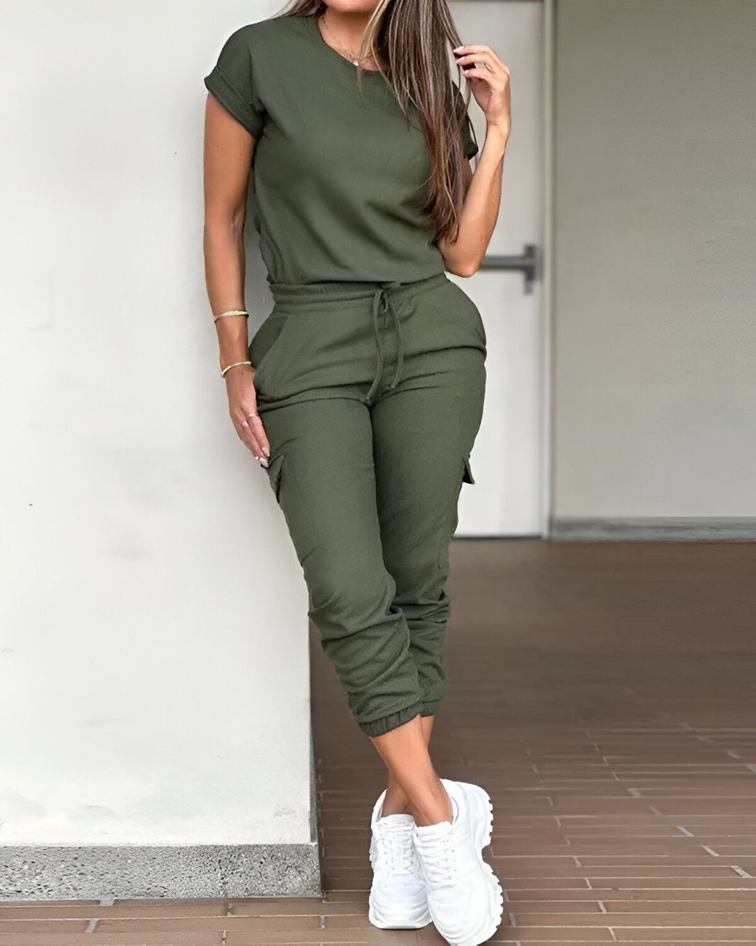 Une femme porte un ensemble de jogging vert kaki composé d'un t-shirt à manches courtes et d'un pantalon cargo. Elle est debout, adossée à un mur, et porte des baskets blanches.
