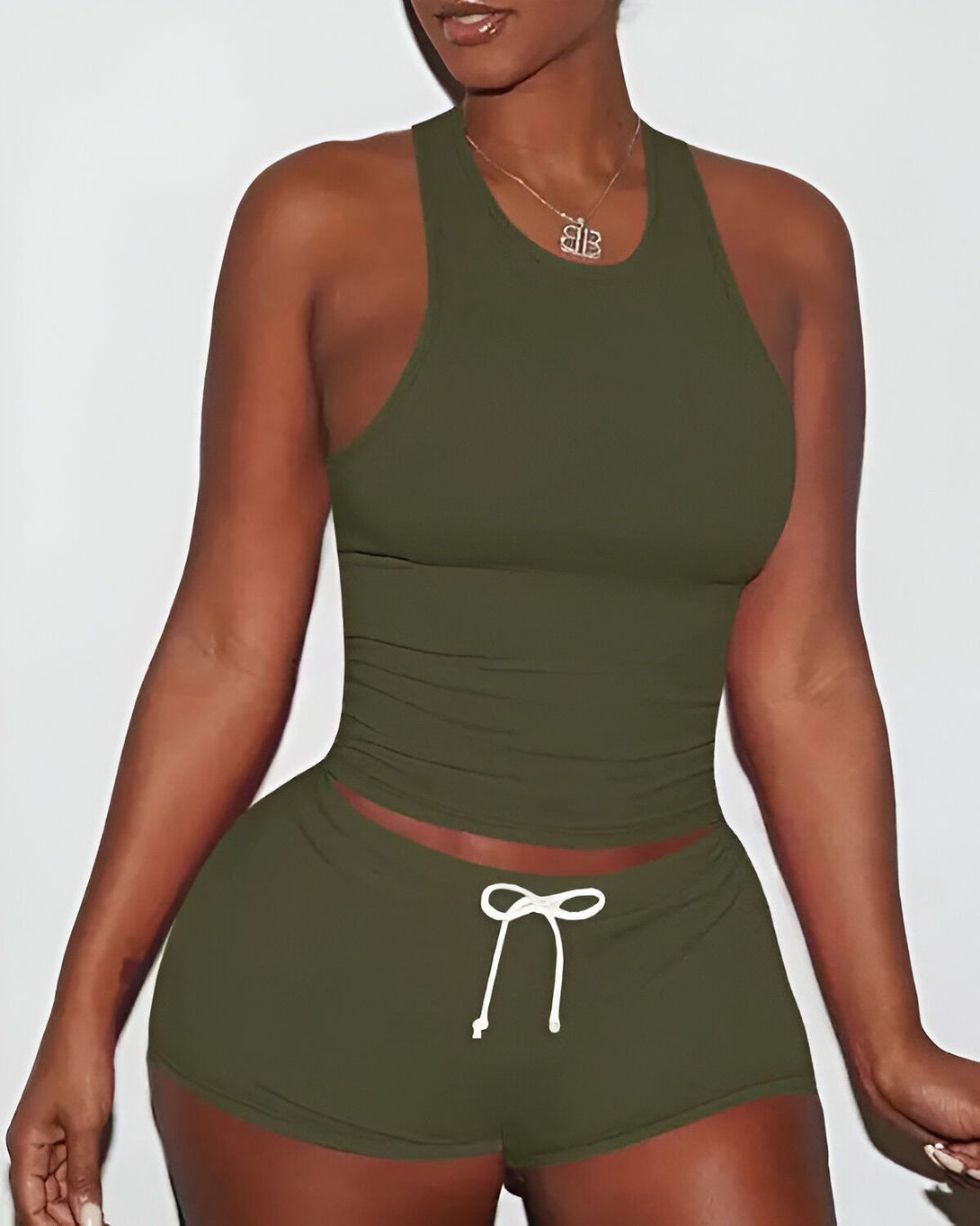 Une femme porte un ensemble de sport vert olive composé d'un débardeur et d'un short. Le débardeur est court et moulant, et le short est taille haute et possède un cordon de serrage à la taille. La femme est vue de face et porte un collier avec un pendentif en forme de B. Elle a les cheveux longs et noirs, et sa peau est mate.