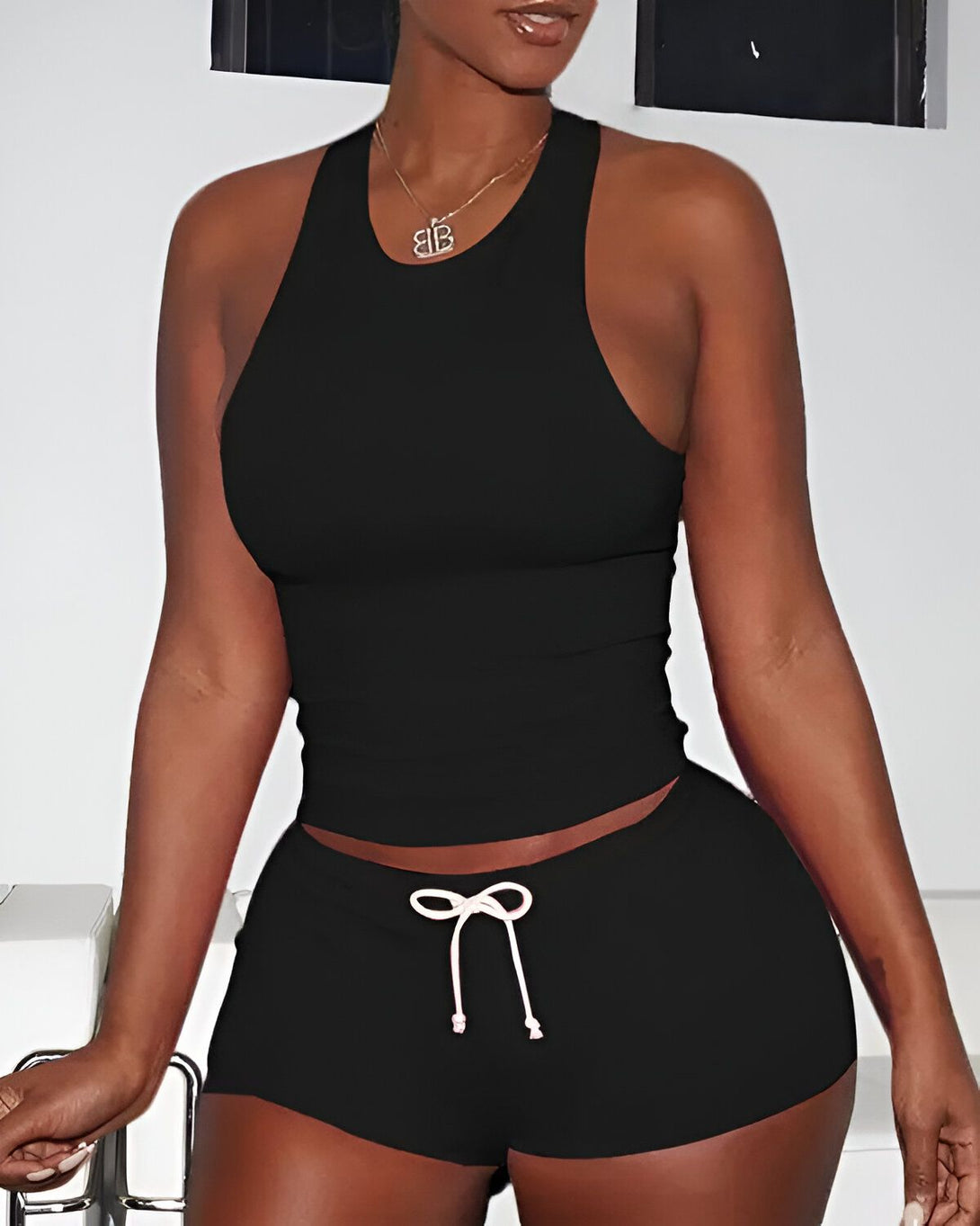 Une femme porte un débardeur et un short noirs. Le débardeur est court et moulant, et le short est taille haute et ample. La femme est debout, de face, et elle regarde l'appareil photo. Elle a la peau foncée et les cheveux noirs. Elle porte un collier avec un pendentif en forme de B.