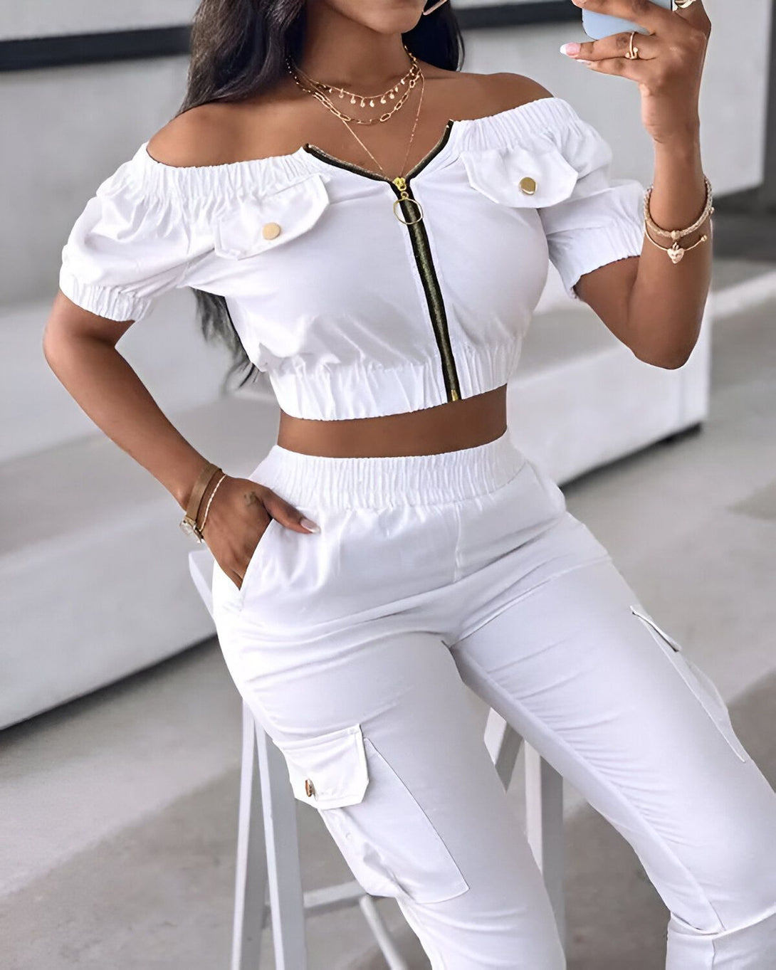 Une femme porte un ensemble deux pièces blanc composé d'un crop top et d'un pantalon cargo. Le crop top est à épaules dénudées et possède une fermeture éclair sur le devant. Le pantalon est taille haute et possède des poches cargo sur les côtés.