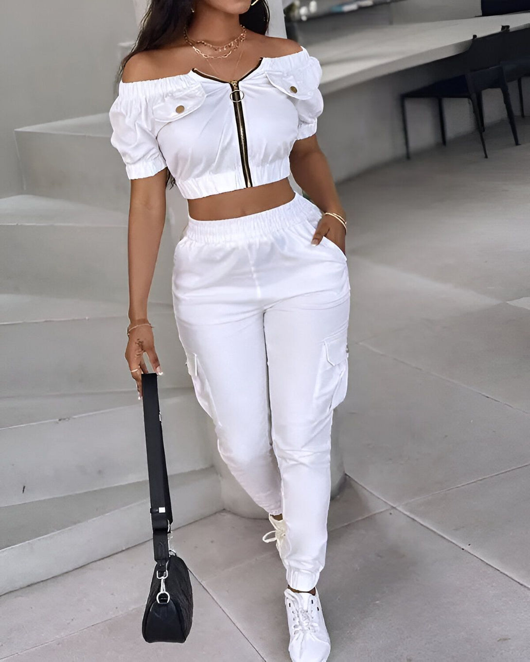 Une femme porte un ensemble deux pièces blanc composé d'un crop top à épaules dénudées et d'un pantalon cargo. Elle porte également des baskets blanches et un sac à main noir.