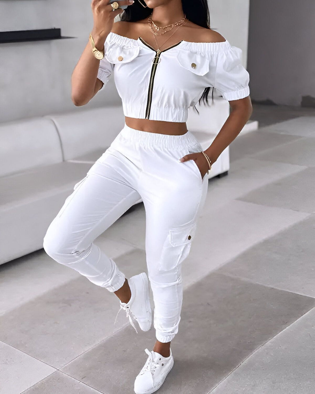 Une femme porte un ensemble de jogging blanc composé d'un haut court à épaules dénudées et d'un pantalon cargo. Elle est également chaussée de baskets blanches.