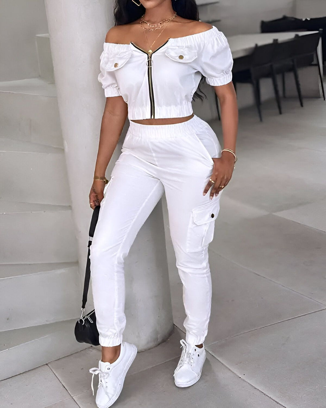 Une femme porte un ensemble pantalon et haut blancs. Le haut est court et épaules dénudées avec une fermeture éclair sur le devant. Le pantalon est cargo avec des poches sur les côtés. La femme porte des baskets blanches et un sac à main noir.