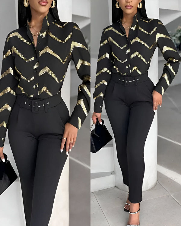 Une femme porte une chemise noire et dorée avec un pantalon noir. Elle est debout et porte des talons noirs. Elle a une main sur la hanche et l'autre tient un sac à main noir.