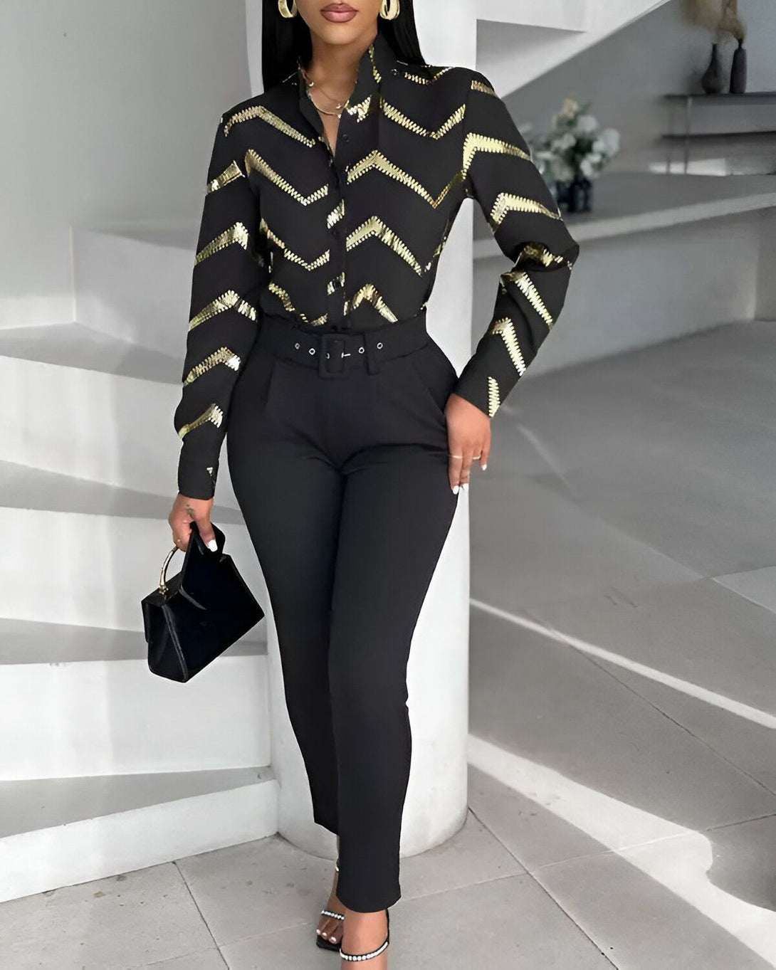 Une femme porte une chemise noire et dorée avec un pantalon noir. Elle porte également des talons noirs et un sac à main noir.