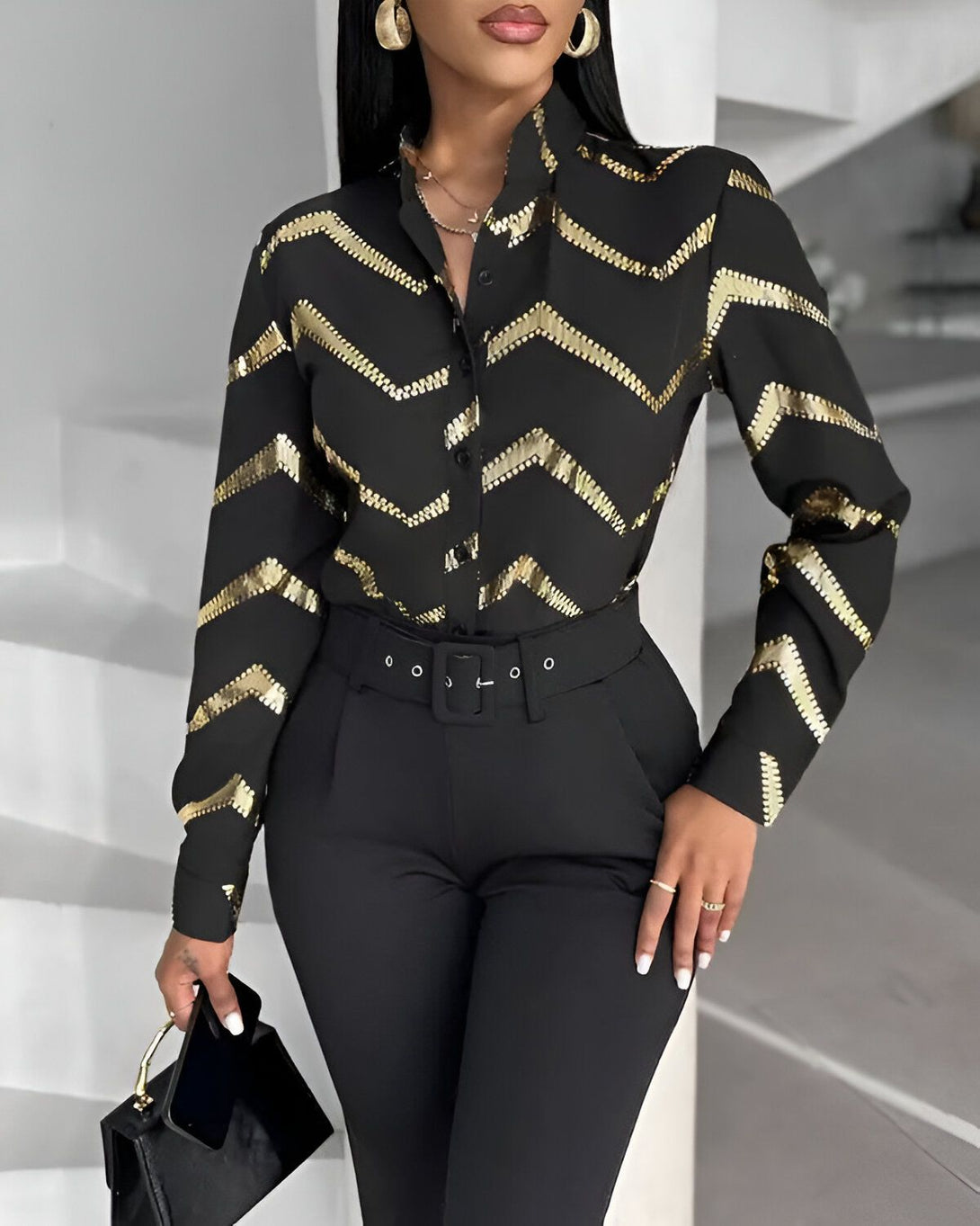 Une femme porte une chemise noire et dorée avec un pantalon noir. Elle est debout dans un escalier et porte une main sur la hanche.