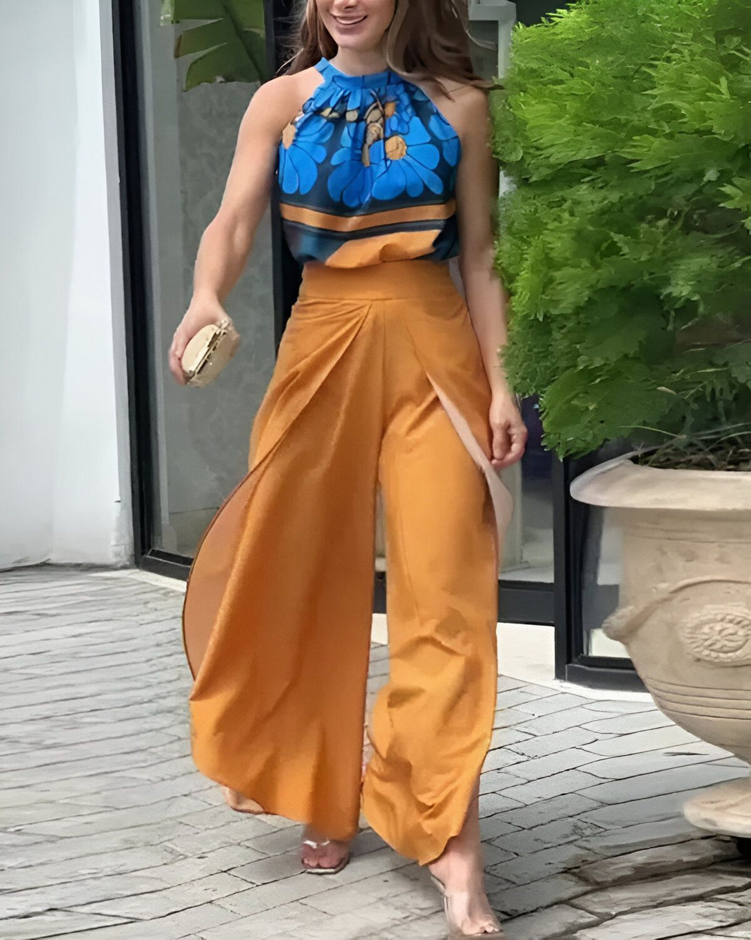 Une femme porte un pantalon orange taille haute et un haut bleu à motifs floraux. Elle est debout devant une porte vitrée.