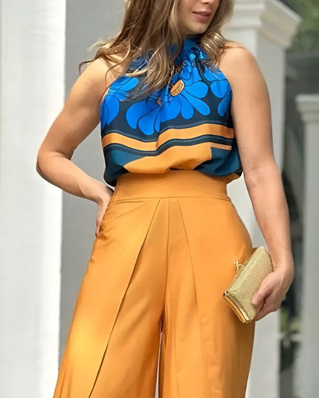 Une femme porte un haut bleu et jaune et un pantalon orange. Elle a un collier et un sac à main.