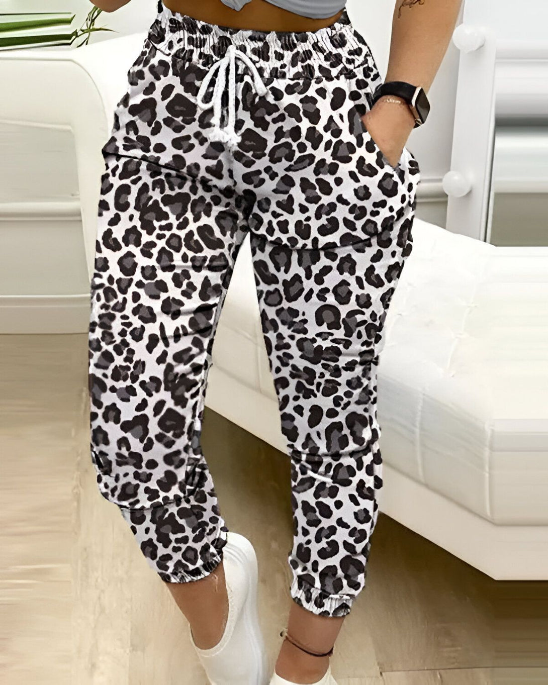 Une femme porte un pantalon de jogging léopard noir et blanc. Le pantalon est taille haute et possède une coupe ample. Il est également doté d'une ceinture à cordon de serrage et de poches latérales. La femme porte un haut blanc et des baskets blanches. Elle a les cheveux longs et bruns et est maquillée de façon naturelle. Elle se tient debout dans un salon et sourit à l'appareil photo.