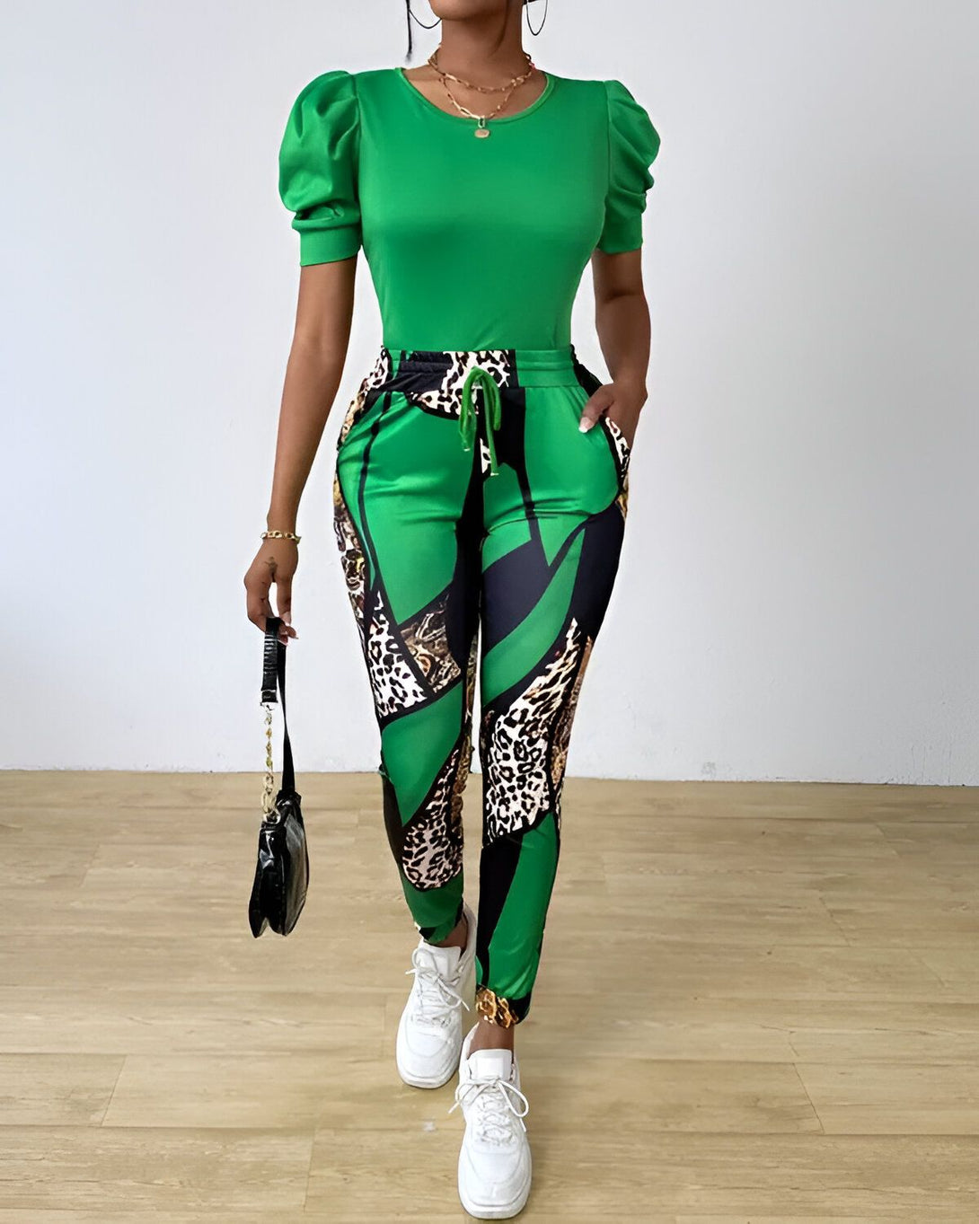 Une femme porte un haut vert à manches bouffantes et un pantalon noir et vert à motifs léopard. Elle porte également des baskets blanches et un sac à main noir.