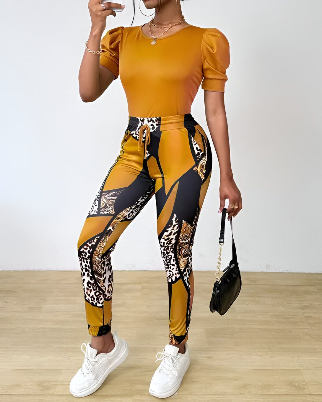 Une femme porte un ensemble pantalon et haut jaune moutarde. Le haut est à manches courtes bouffantes et le pantalon est taille haute et possède des imprimés animaliers. Elle porte des baskets blanches et un sac à main noir.