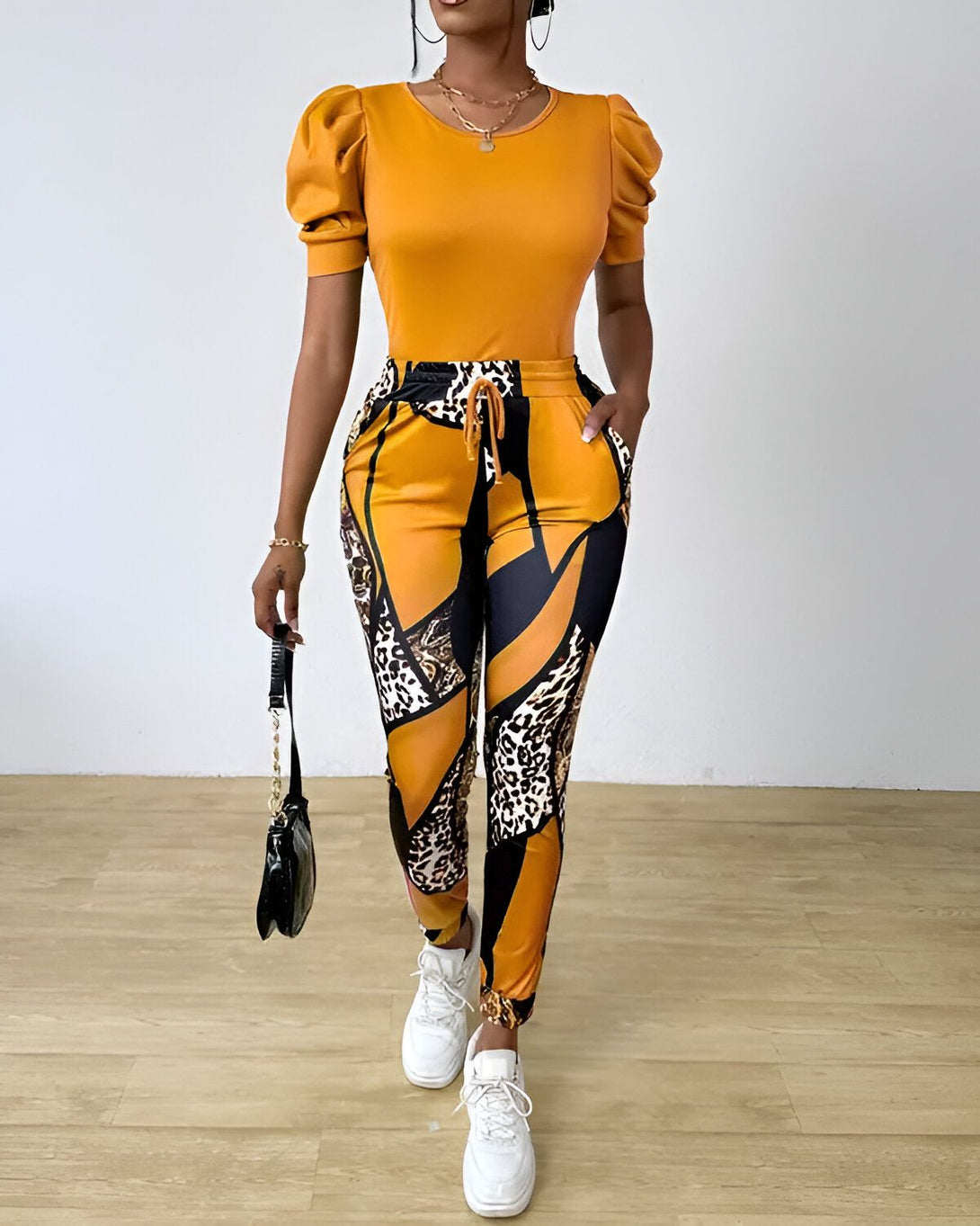 Une femme porte un ensemble pantalon et haut jaune et noir. Le haut est à manches courtes et bouffantes. Le pantalon est taille haute et possède une ceinture à nouer. La femme porte également des baskets blanches et un sac à main noir.