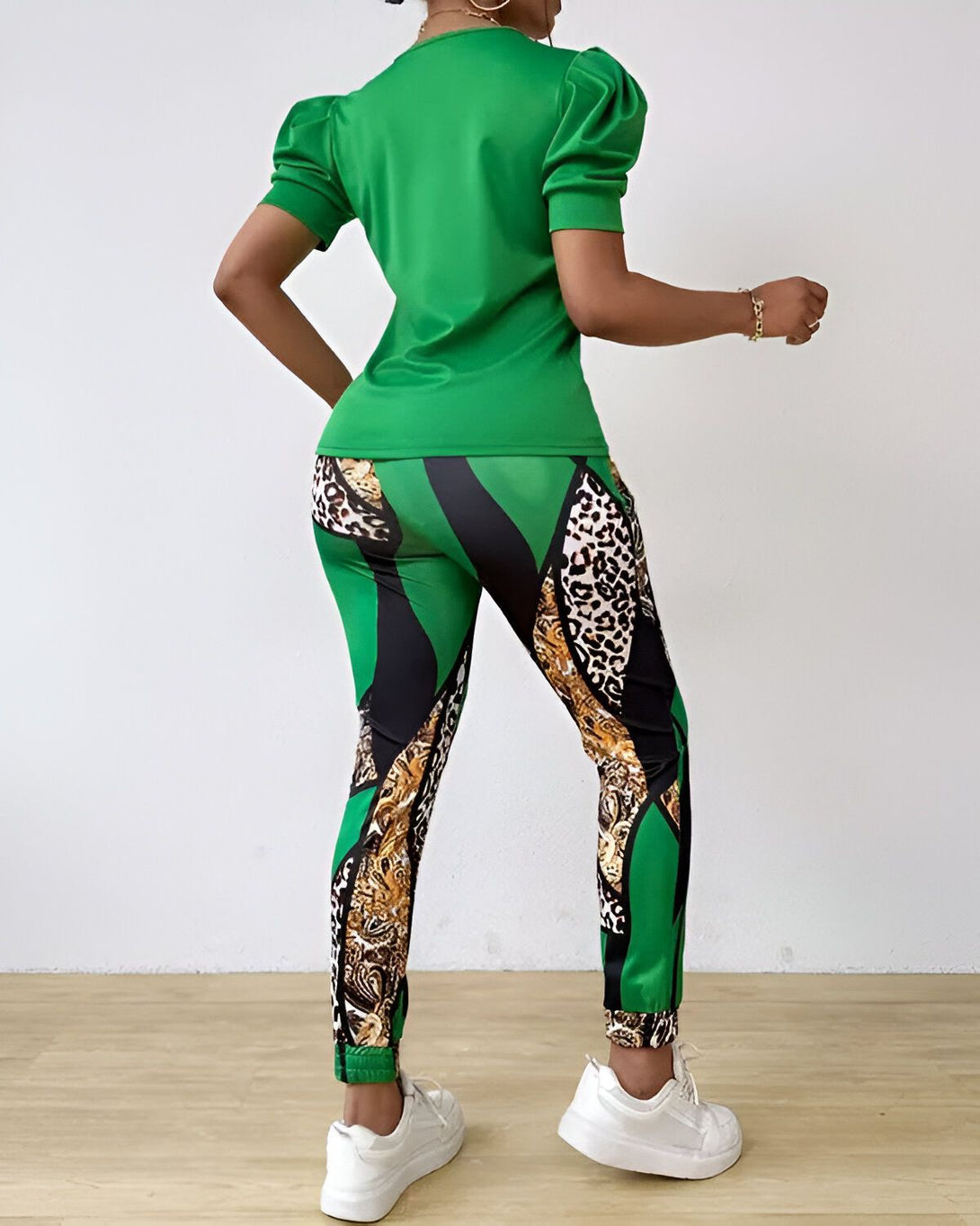 Une femme porte un ensemble vert et noir. Le haut est vert avec des manches bouffantes et un col rond. Le pantalon est noir avec des motifs léopard et des rayures vertes. La femme est vue de dos et porte des baskets blanches.