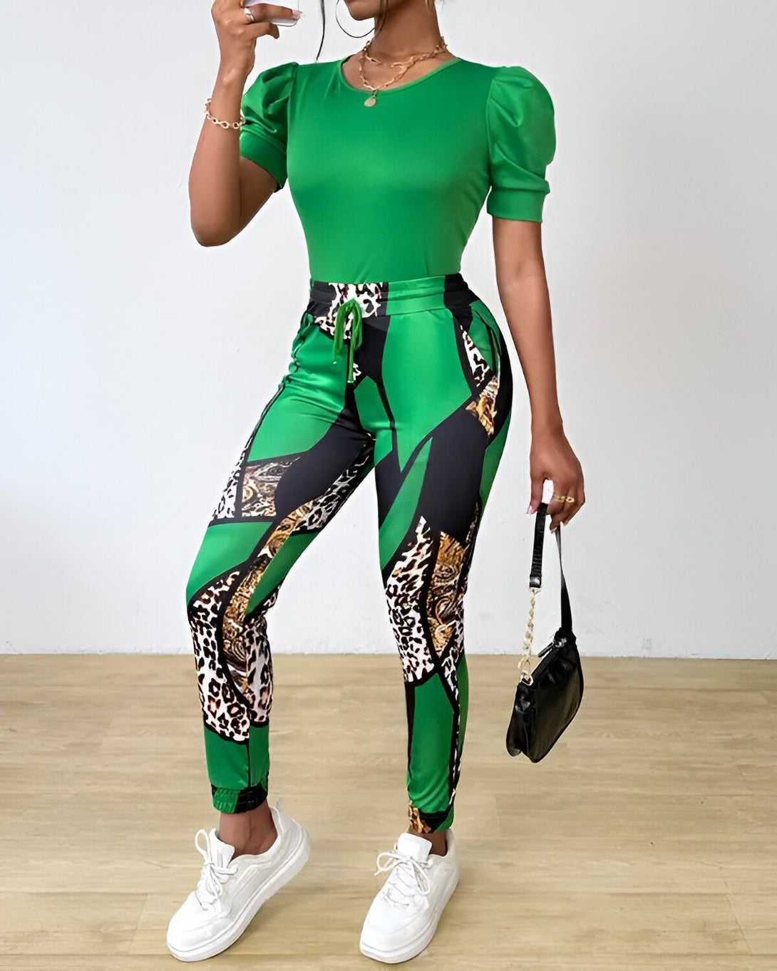 Une femme porte un ensemble vert composé d'un haut à manches bouffantes et d'un pantalon. Le haut est vert uni, tandis que le pantalon est imprimé de motifs léopard et de chaînes. La femme porte également des baskets blanches et un sac à main noir.