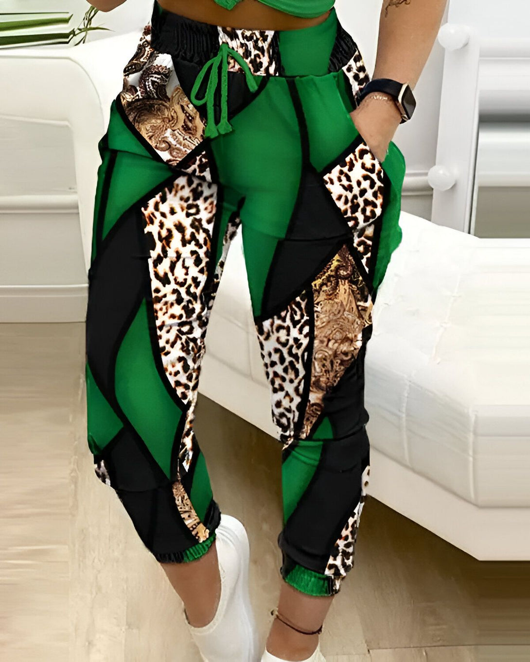 Une femme porte un pantalon de jogging vert et noir. Le pantalon est imprimé de motifs léopard et géométriques. La femme est vue de face et elle a les mains dans les poches.