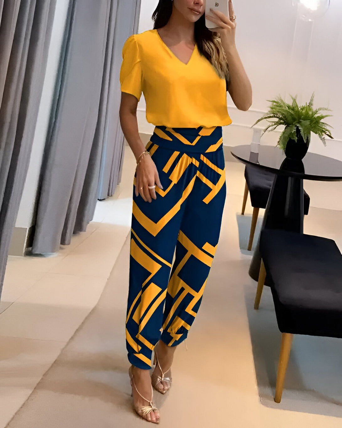 Une femme porte un chemisier jaune à manches courtes et un pantalon bleu à motifs géométriques jaunes. Elle est debout dans un salon, une main sur la hanche et l'autre tenant un téléphone portable.
