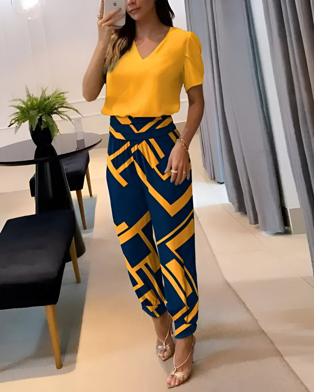 Une femme porte un chemisier jaune à manches courtes et un pantalon bleu marine à motifs géométriques jaunes. Elle est debout dans un salon, près d'une table et d'une chaise.