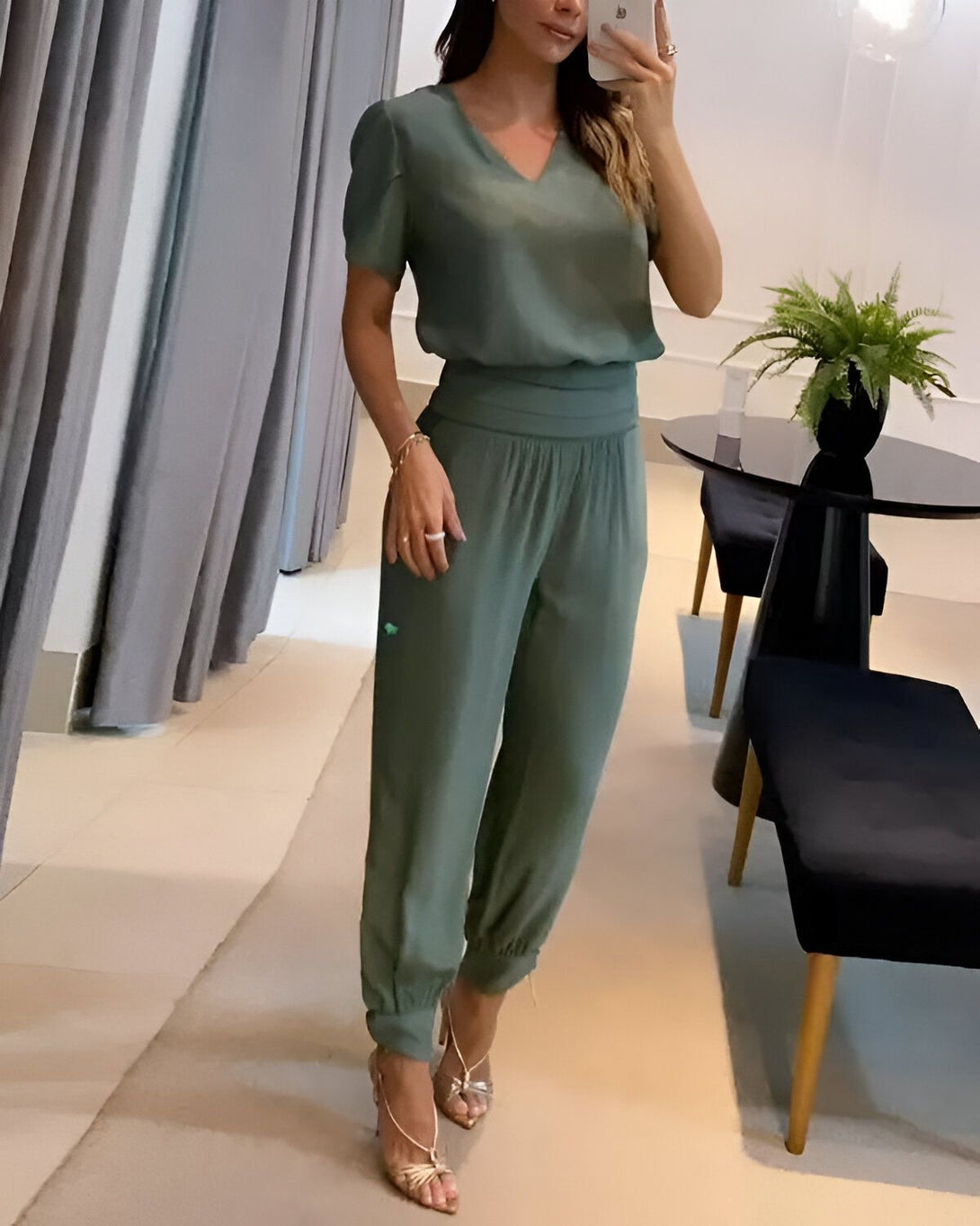 Une femme porte un pantalon et un chemisier vert olive. Elle est debout dans un magasin et se prend en photo dans un miroir.