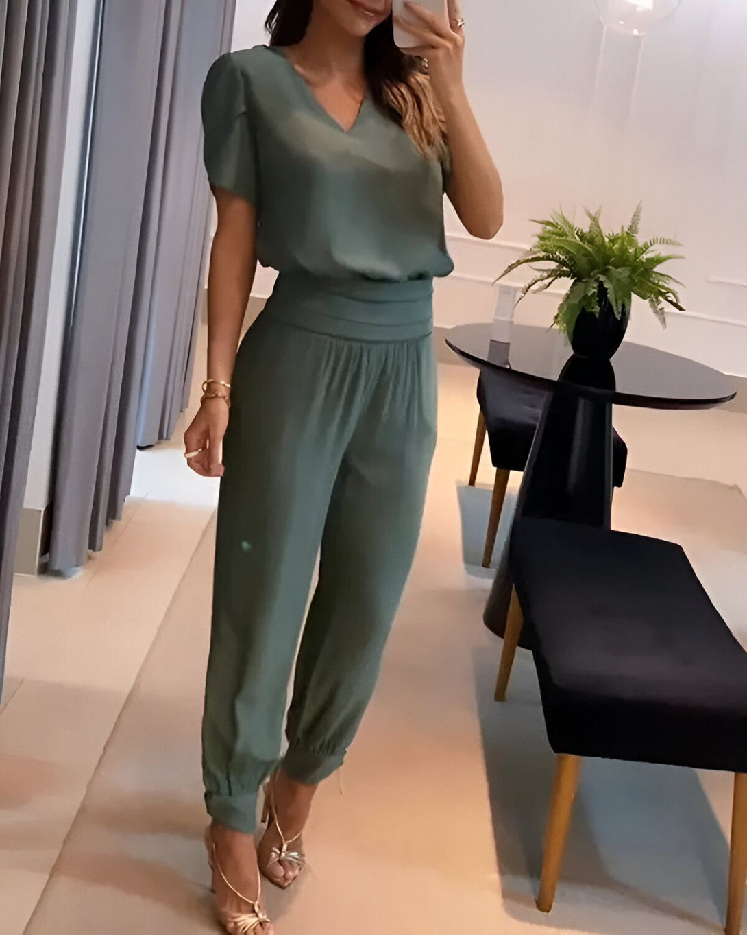 Une femme est debout dans un salon. Elle porte un chemisier vert à manches courtes et un pantalon vert assorti. Elle porte également des sandales à talons dorées et un bracelet doré au poignet gauche. Ses cheveux sont longs et bruns et elle porte un maquillage léger.
