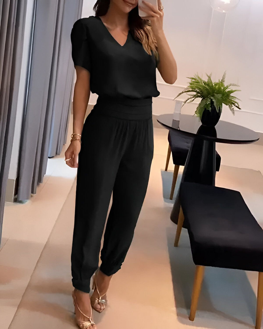 Une femme porte un ensemble pantalon et haut noir. Le haut est à manches courtes et le pantalon est taille haute. Elle porte des sandales à talons hauts dorées et un bracelet doré au poignet gauche.