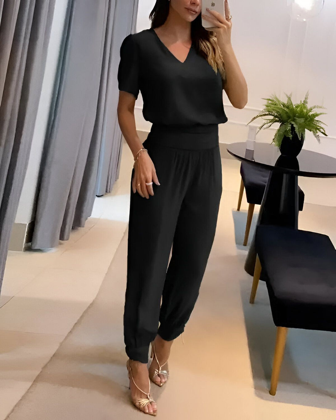 Une femme est debout dans un salon. Elle porte un chemisier noir à manches courtes et un pantalon noir taille haute. Elle est chaussée de sandales à talons hauts dorées. Ses cheveux sont longs et bruns. Elle est en train de se prendre en photo avec son téléphone portable.