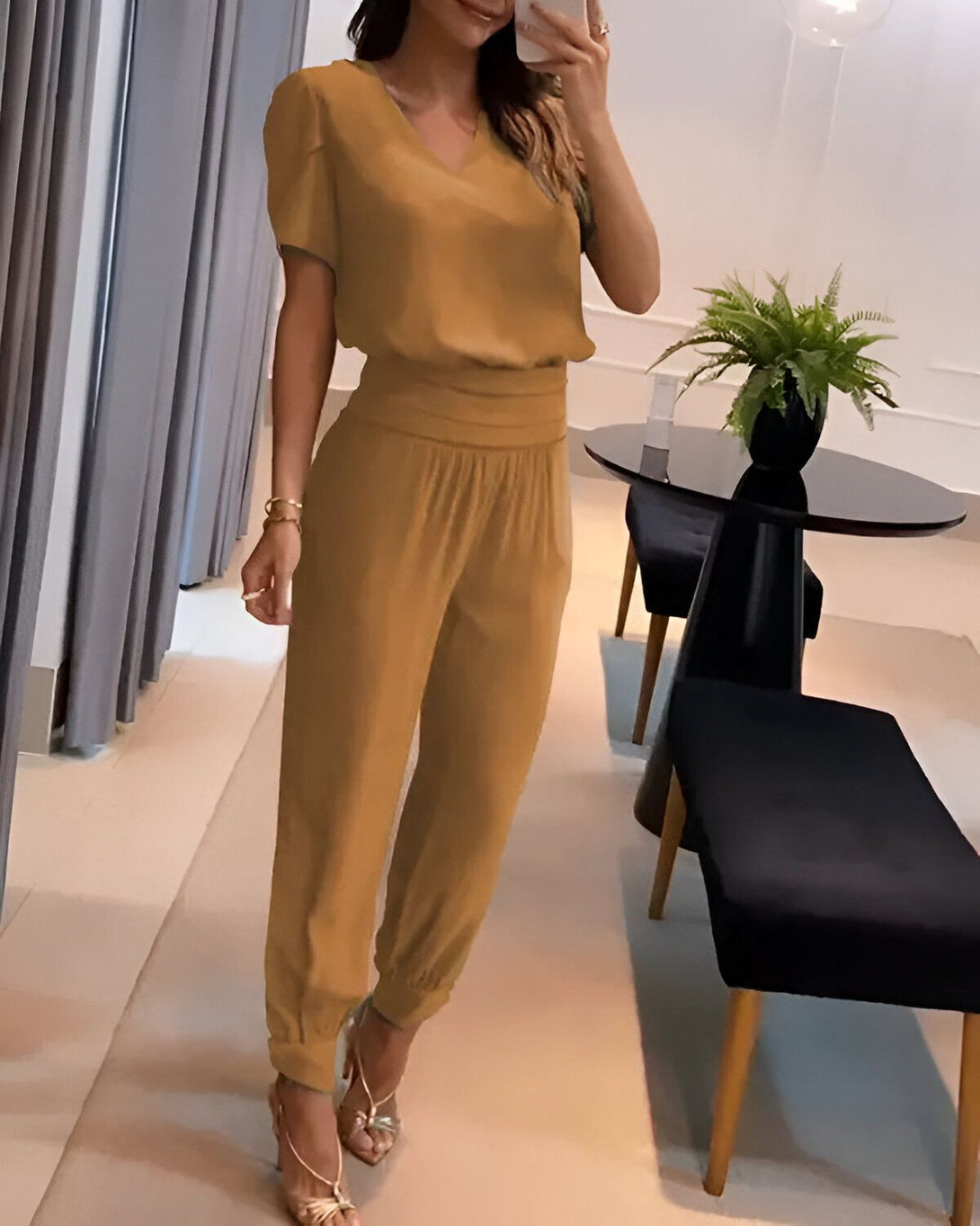 Une femme porte un ensemble pantalon et haut marron. Le haut est à manches courtes et le pantalon est taille haute. Elle porte des sandales à talons hauts et un collier.