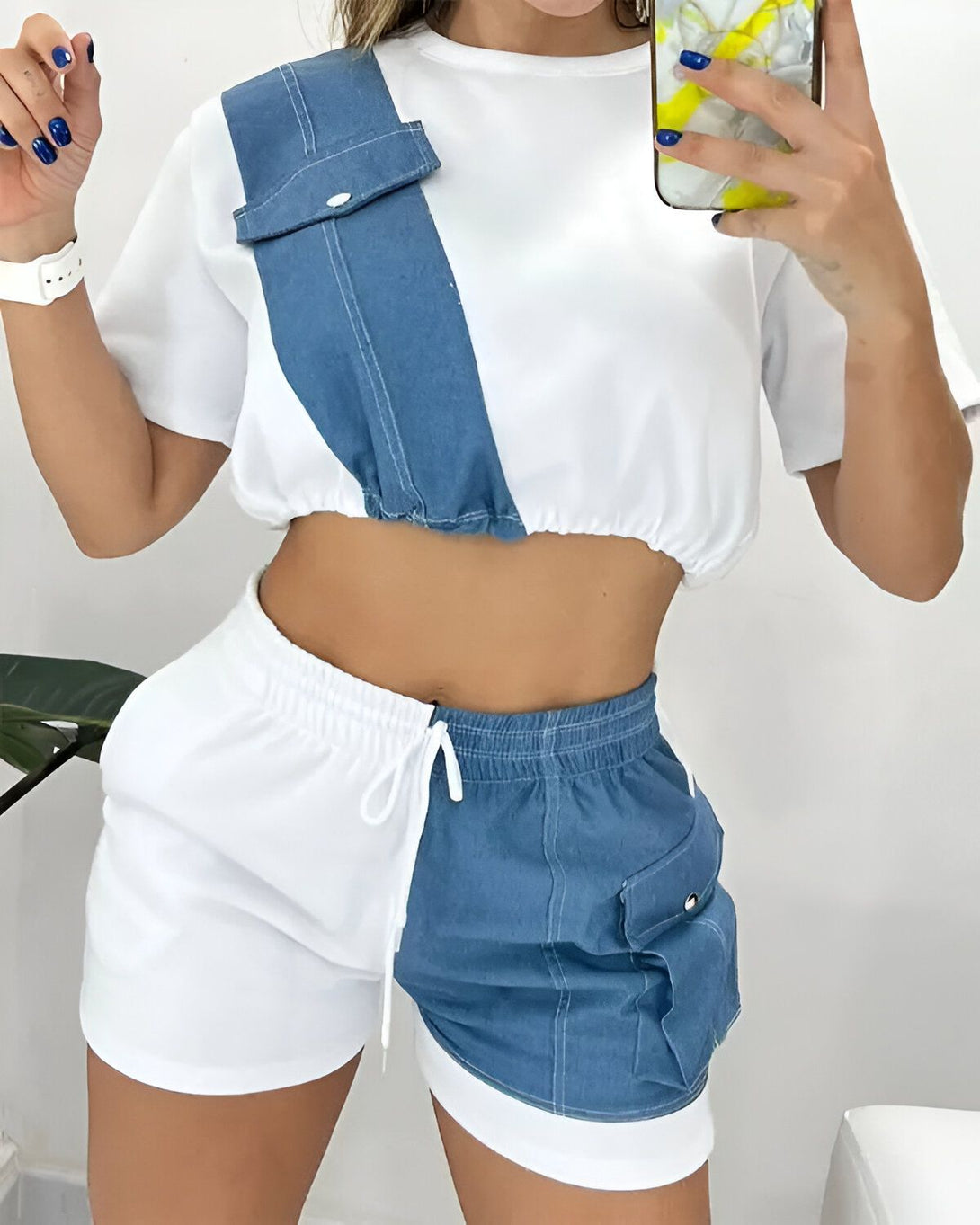 Une femme porte un ensemble deux pièces composé d'un crop top et d'un short. Le crop top est blanc avec une poche en jean sur le devant. Le short est blanc et bleu marine, avec une taille haute et des poches cargo. La femme est également porte une montre blanche au poignet gauche et tient un téléphone portable dans sa main droite.