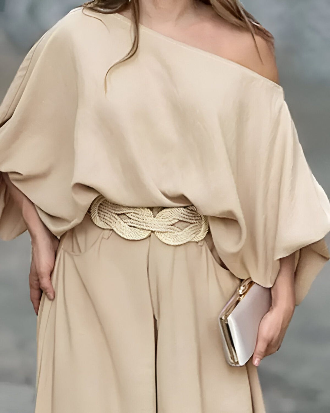 Une femme porte une combinaison pantalon beige avec une ceinture tressée dorée et un sac à main argenté.