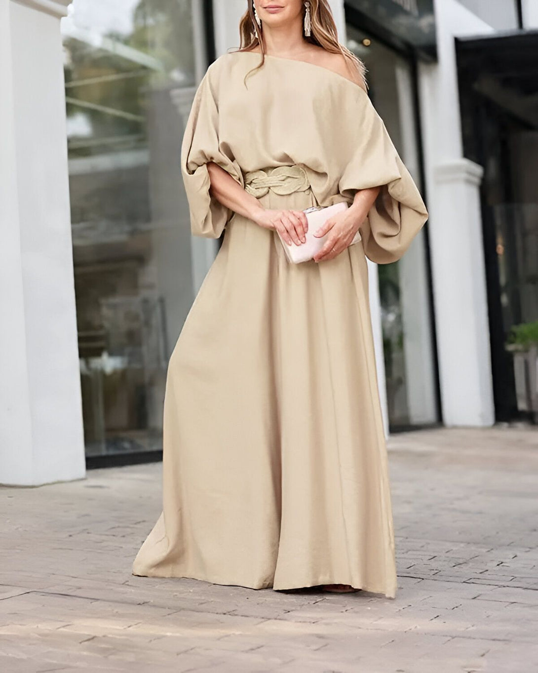Une femme porte une robe longue beige avec une manche bouffante et une ceinture. Elle porte également des chaussures à talons beiges et un sac à main rose.