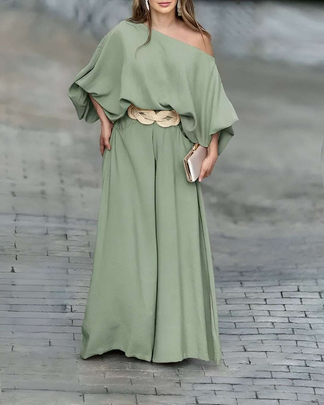 Une femme porte une combinaison pantalon vert olive à une épaule et une ceinture tressée dorée. Elle porte également un sac à main doré.