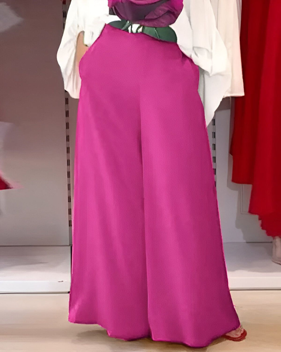 Une femme porte un pantalon large rose fuchsia. Le pantalon est taille haute et a des poches. La femme porte également un chemisier blanc et des sandales roses.