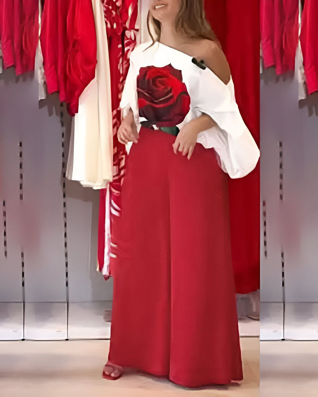 Une femme porte un chemisier blanc à épaules dénudées avec une rose rouge imprimée sur le devant et un pantalon rouge assorti. Elle porte des sandales rouges à talons hauts et se tient dans une boutique de vêtements.