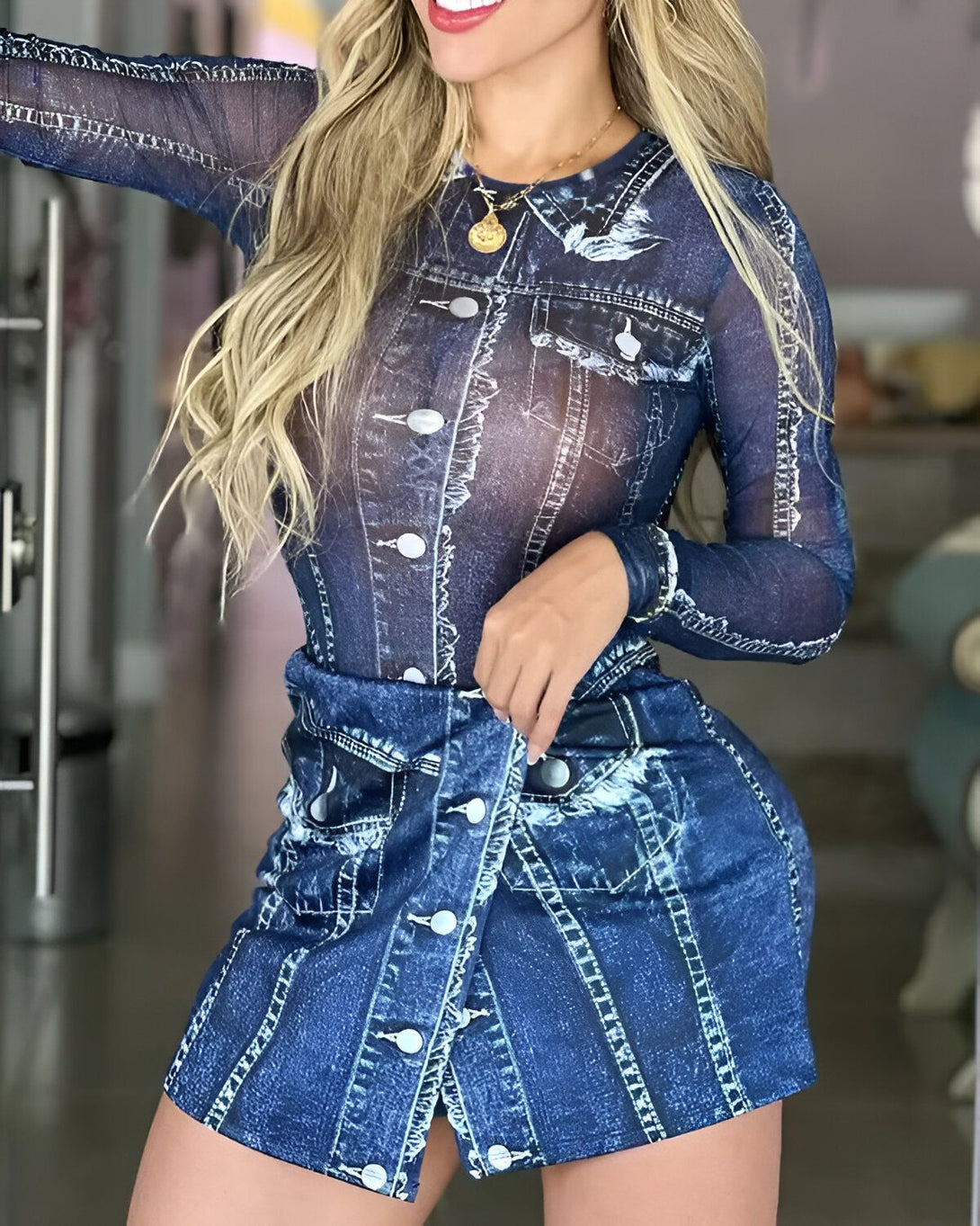 Une femme porte un ensemble jupe et haut en jean. Le haut est transparent et laisse entrevoir sa poitrine. La jupe est courte et taille haute. Elle est boutonnée sur le devant. La femme est blonde et a les cheveux longs. Elle porte un collier et des boucles d'oreilles. Elle est souriante et regarde l'objectif.