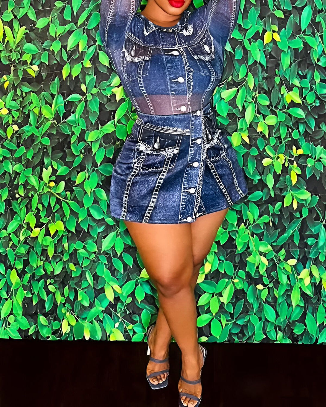 Une femme porte une robe en jean bleu avec des talons transparents. La robe est courte et cintrée à la taille. La femme a les cheveux longs et noirs et porte du rouge à lèvres rouge. Elle pose devant un mur de feuilles vertes.